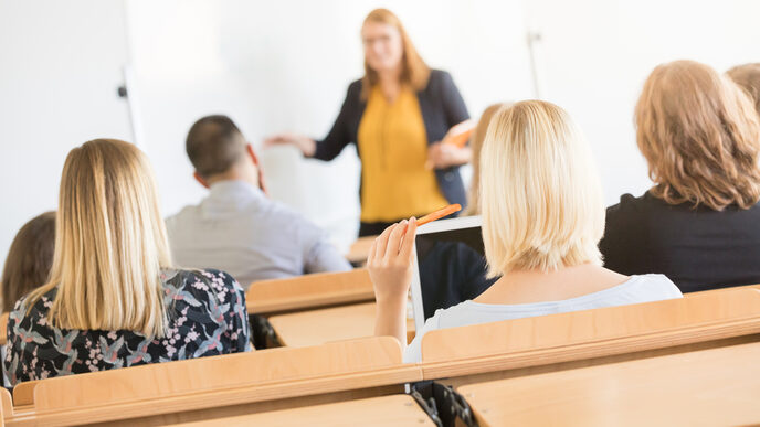 Foto von Hinterköpfen mehrerer Studierender in Sitzreihen im Hörsaal. Vorne ist  eine Dozentin unscharf zu erkennen.