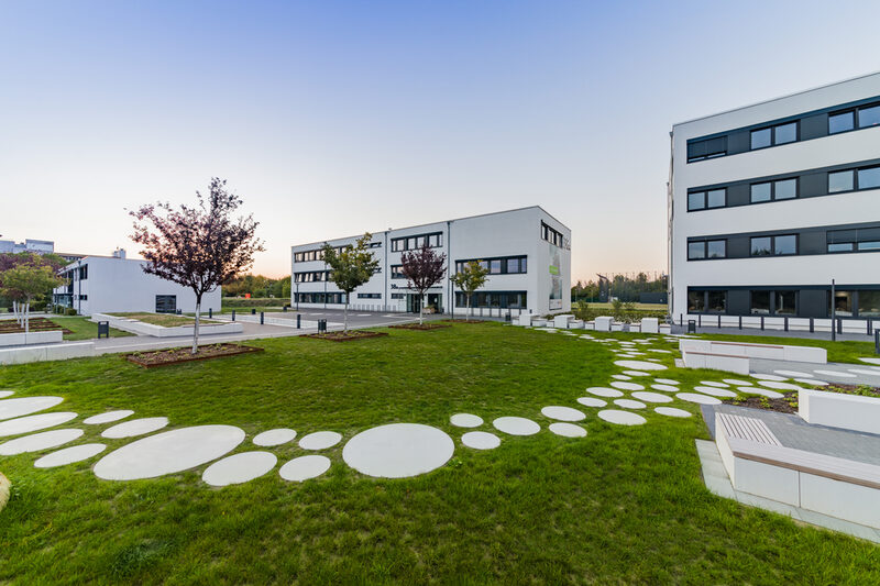 Foto des Gebäudes 38 a der Emil-Figge-Straße, daneben links das Gebäude 38 im Hintergrund und rechts im Anschnitt das Gebäude 38 b. Im Vordergrund ist die Wiese mit Bäumen sowie runde Bodenplatten als Gehweg. __ <br>View of building 38 a on street Emil-Figge, next to it you can see building 38 in the background on the left and building 38 b on the right. In the foreground you can see the meadow with trees and the round floor slabs on the meadow that lead to building 38 b.