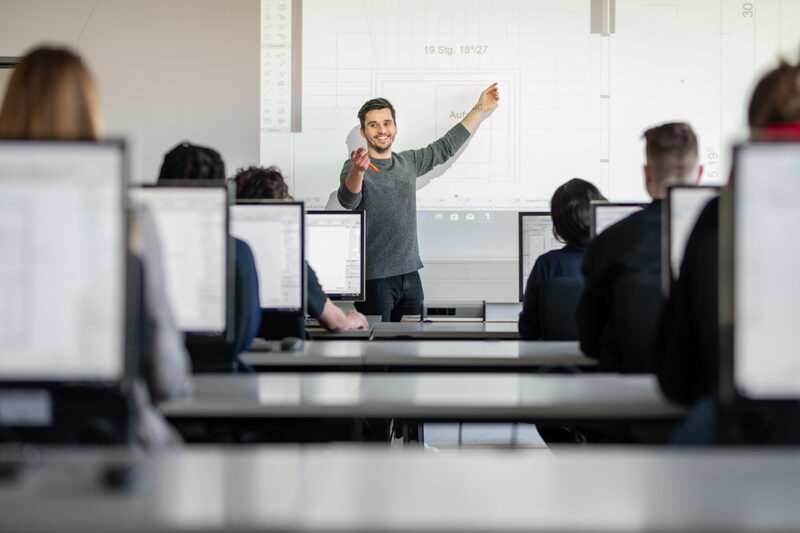 Foto eines Dozenten im Computerraum. Er zeigt etwas an einer Grafik, die auf die Wand projiziert wurde. Mit der anderen Hand zeigt er lächeln zu einem Studierenden, als würde er diese Person aufrufen. __ <br>An assistant professor is standing in the front of the computer room and is showing something on a graphic that has been projected onto the wall. With the other hand he shows a student smile as if he were calling that person.