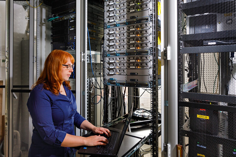 Photo of a trainee stands in front of a large cabinet with servers and operates a laptop. __ <br>A trainee stands in front of a large cabinet with servers and operates a laptop.