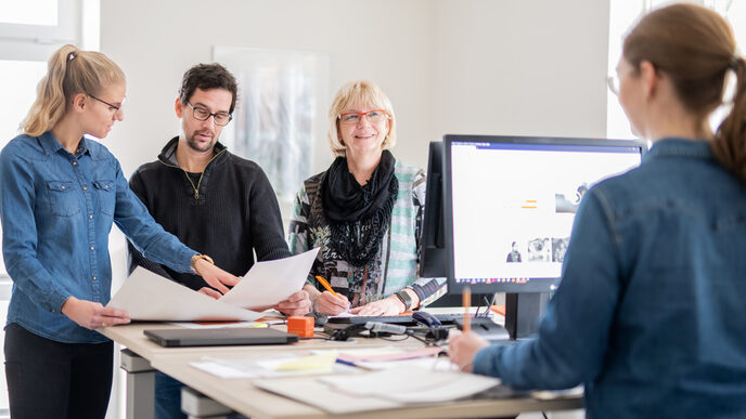 Photo of four employees in the administration department at their desks, one of them seen from behind. This person is talking to a woman opposite her, the other two employees are looking through documents.