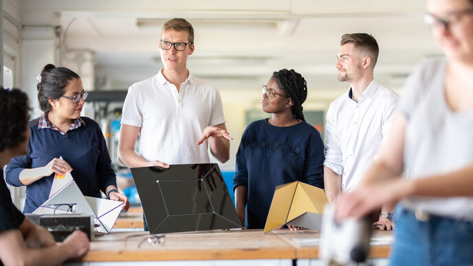 Photo of a lab employee holding a metal object in his hands that is to be folded and explaining something. Other students are standing around him and the workbench, watching the two of them. In the foreground, out of focus, is another student working on something.