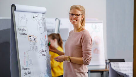 Foto einer Mitarbeiterin bei einem Seminar der Personalentwicklung. Sie steht vor einem Flipchart, auf dem sich mehrere bunte Zeichnungen und Beschriftigungen befinden.