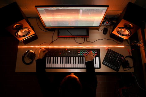 Seen from above: A person is sitting at a desk with a keyboard, a monitor, two loudspeakers, headphones and other sound equipment.