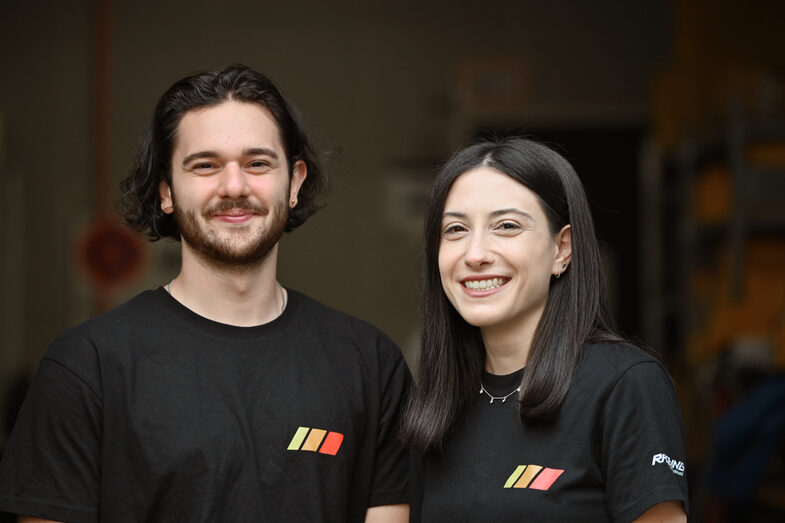 Two people in black T-shirts in a portrait in front of a dark room in the background.