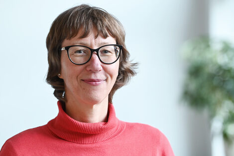 A person in a portrait with a red sweater in front of a light gray background, looking into the camera and smiling.