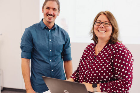 Two people stand at a desk with a laptop and look into the camera with a smile.