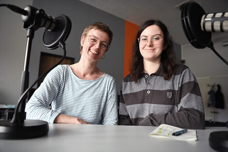 Two people sit at a table between two microphones and look smiling into the camera.