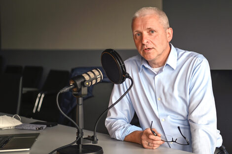 A man in a blue shirt is sitting at a table with a microphone, talking and looking into the camera, holding his glasses in his right hand.