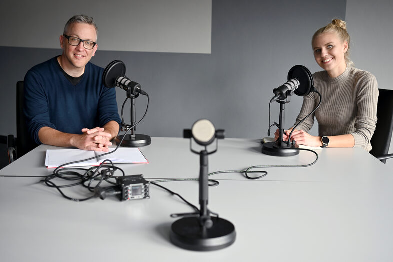 A woman (right) and a man (left) are sitting at a table facing the viewers. There is a table microphone in front of each of them. A third microphone is in the middle, facing the viewers.