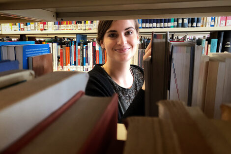 Eine Frau schaut durch ein Bücherregal hindurch in die Kamera.