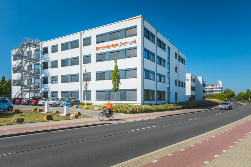 Foto eines Gebäudes der FH Dortmund in der Emil-Figge-Straße 38b mit Sicht auf das leuchtende FH-Logo und Straße und Parkplatz im Vordergrund.