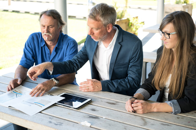 Photo of three employees sitting at a table outside and looking at documents together.