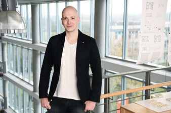 Ein Mensch im schwarzen Sakko steht in einem Gebäude an einem Geländer vor einer Fensterfront.