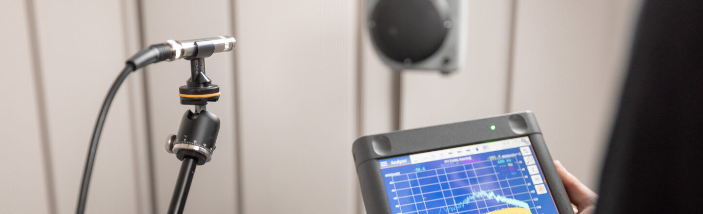 Ein Messmikrofon steht in einem Schallmessraum vor einem Lautsprecher. Eine Person hält die Messtechnik in den Händen auf der die Schallmessung grafisch dargestellt ist.