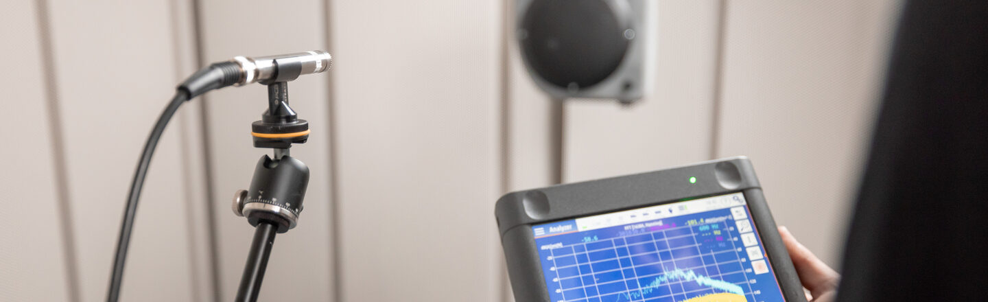 A measuring microphone is placed in a sound measurement room in front of a loudspeaker. One person is holding the measuring equipment in their hands on which the sound measurement is graphically displayed.