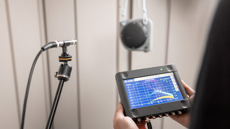 A measuring microphone is placed in a sound measurement room in front of a loudspeaker. One person is holding the measuring equipment in their hands on which the sound measurement is graphically displayed.