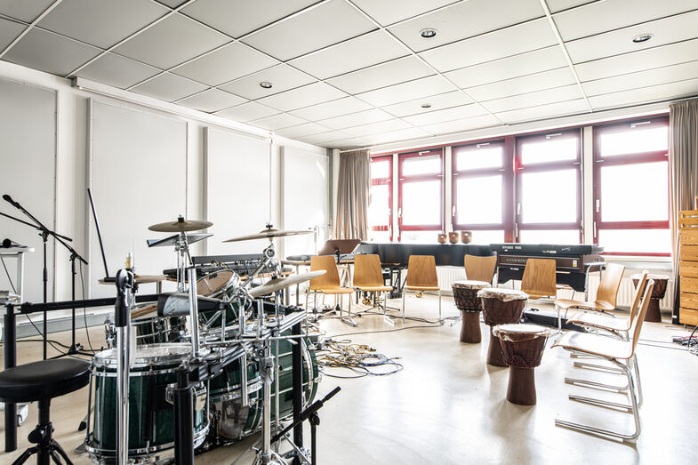 Foto des Audiolabors. Links im Bild ein Schlagzeug und Mikrofonständer. Rechts im Bild Stühle und drei Djembe-Trommeln, im hinteren Teil des Raumes ein Flügel.
