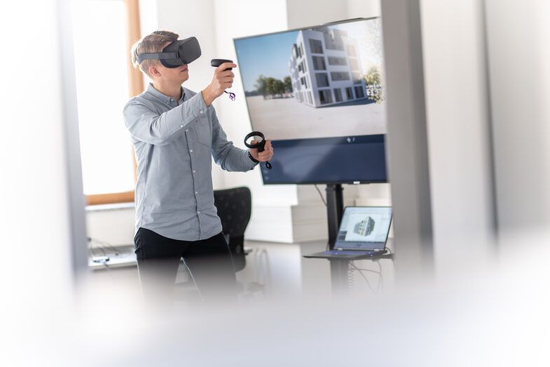 Foto von einem Mann mit VR-Brille auf und Controller in der Hand. Auf dem Bildschirm im Hintergrund sieht man ein Gebäude in der Perspektive, die auch der Mann durch die VR-Brille sieht.
