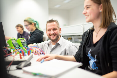 Foto eines Professors, der im Labor für Elektronik und Automation neben einer Studentin hockt, die an einem Computer arbeitet, beide schauen auf den PC-Bildschirm. Auf dem Tisch stehen zudem zwei Messgeräte mit diversen Kabeln. Am Tisch daneben arbeiten zwei weitere Personen.