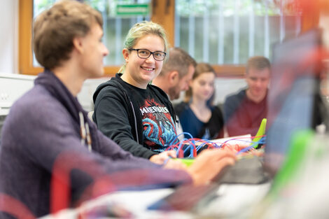 Foto von mehreren Studierenden, die nebeneinander an einer Tischreihe mit verschiedenen elektronische Versuchsaufbauten sitzen. Eine Studentin ist im Fokus und lächelt in die Kamera.