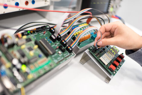 Foto einer männlichen Hand, die ein Kabel in einen elektrotechnischen Aufbau mit Platinen steckt.<br>Auf dem vorderen Aufbau sind Zahlen auf einem Feld.