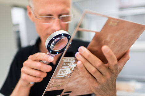 Foto eines Mitarbeiters des Labors für Elektronik und Automation, der mit Hilfe einer Lupe eine gefräste Platine begutachtet.