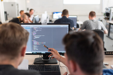 Photo of students in rear view at computer workstations. In the foreground, a student shows something on the screen to the person sitting next to him with a pen.