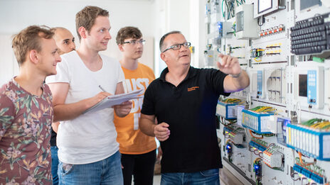 Photo of a professor with his students. They are standing in front of an electrical engineering test wall, the professor is pointing at a device. One of the students is taking notes.