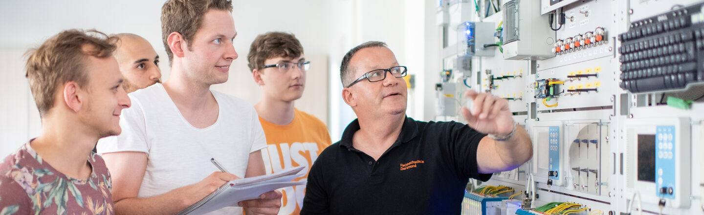 Photo of a professor with his students. They are standing in front of an electrical engineering test wall, the professor is pointing at a device. One of the students is taking notes.