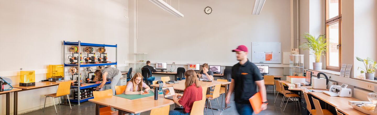 Raumaufnahme von einem Werkraum in dem verschiedene Arbeitsplätze mit verschiedenster Technik ist, z.B. 3D-Drucker, Computer, Laser, Plotter, Nähplatz. Im Raum arbeiten Studierende an verschiedenen Stationen.