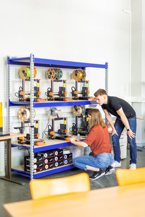 Photo of two students standing/squatting at the 3D printer shelf.