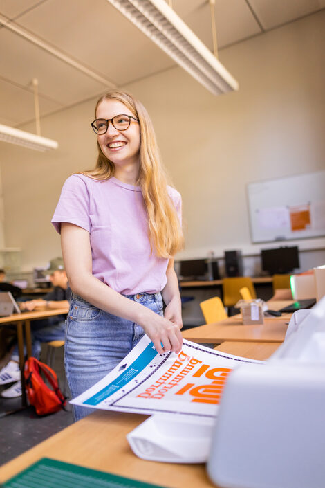 Foto von einem Plotter, davor steht eine Studentin, die die Plotterarbeit entgegen nimmt.