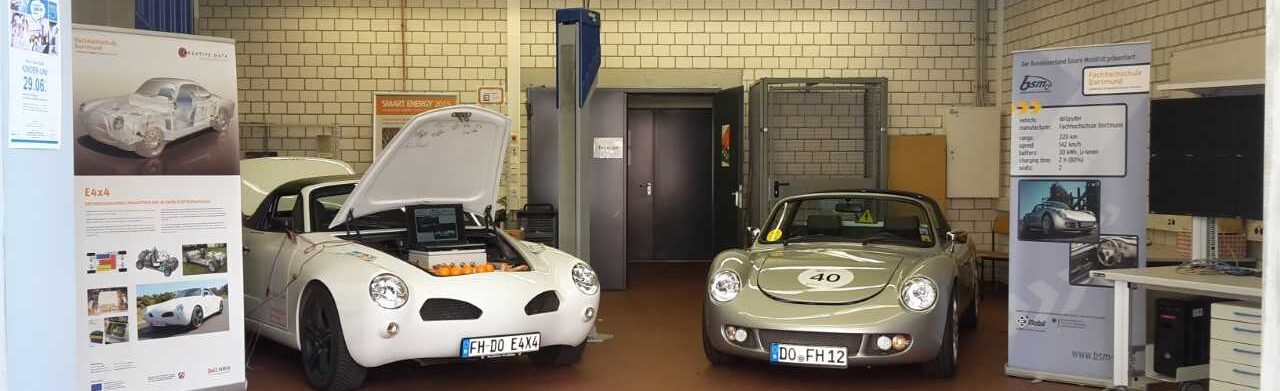 Foto von zwei Autos in einer Werkstatt. Bei einem der Autos ist die Motorhaube geöffnet. In der Werkstatt befinden sich Aufsteller mit technischen Informationen zu den Autos.