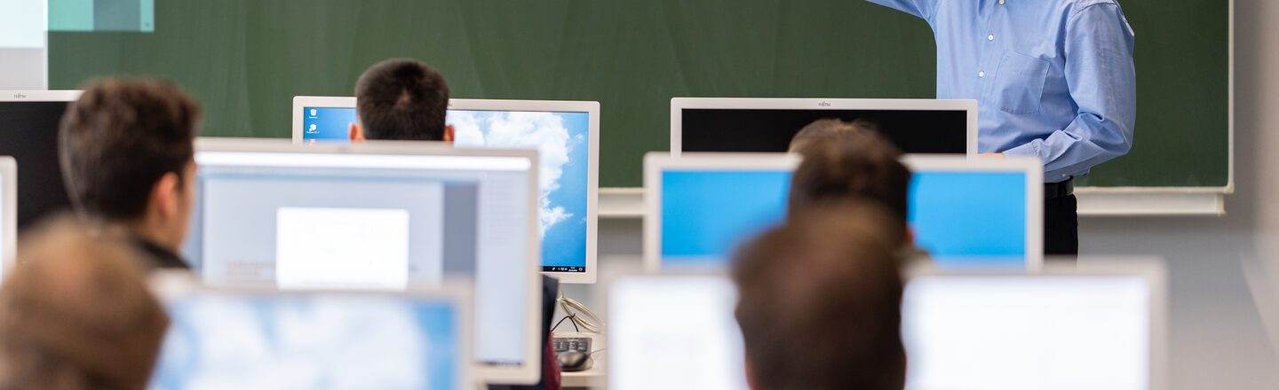 Photo of a professor showing something on the blackboard in the computer room. In front of him are students at computers.