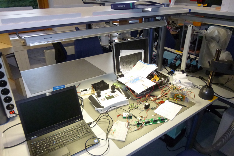 Foto eines Tisches mit einem Laptop, vielen Kabeln und diversen technischen Geräten, wie zum Beispiel einer digitalen Fernsteuerung.