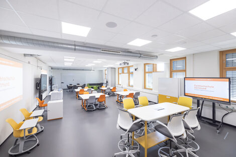 Image of the InnoLab, a modular and divisible room with various work areas. In the foreground is a high table with chairs, on the sides several digital whiteboards, a lectern and a projector surface as well as group tables. At the back there is a couch and a meeting table as well as walls to divide the room.