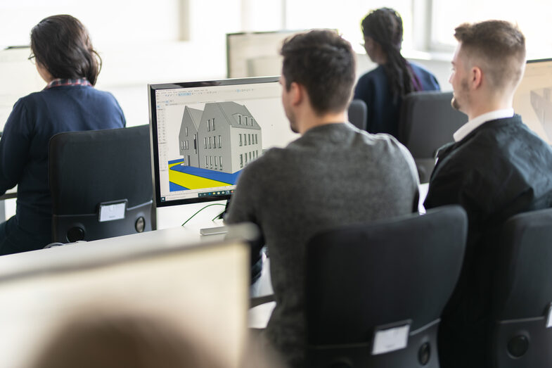 Foto mehrerer Studierender in Rückansicht, die an PCs arbeiten. Auf einem Bildschirm zweier Studenten ist ein Entwurf eines Hauses zu erkennen.