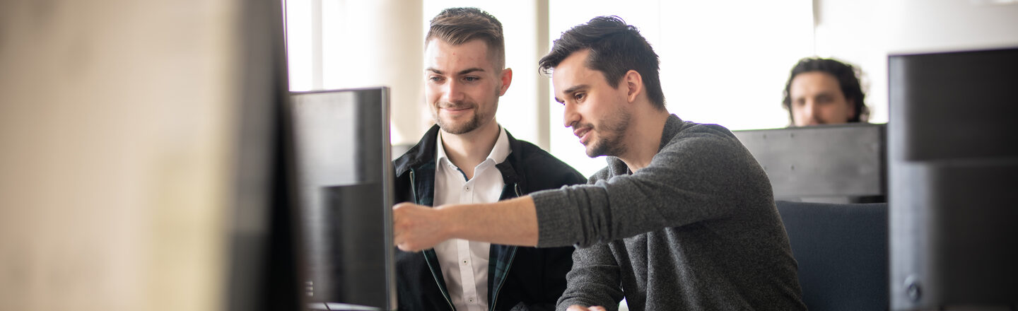 Foto eines Dozenten neben einem Studenten am PC. Der Dozent zeigt dem Studenten etwas auf dem Bildschirm. Im Hintergrund ein weiterer Student an einem Computer.