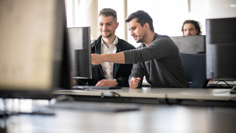 Foto eines Dozenten neben einem Studenten am PC. Der Dozent zeigt dem Studenten etwas auf dem Bildschirm. Im Hintergrund ein weiterer Student an einem Computer.