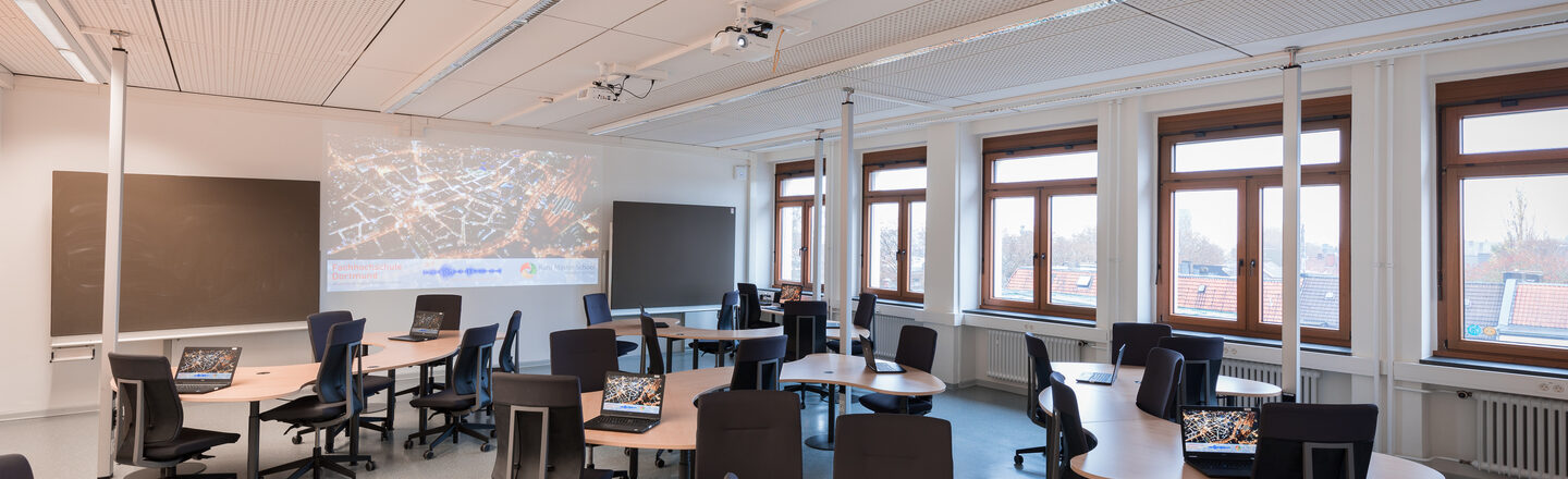 Room shot of the ICT lab with activated laptops on the tables. The tables have an unusual curved shape. A projection can be seen on the wall.