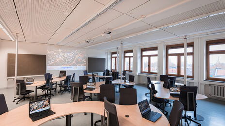 Room shot of the ICT lab with activated laptops on the tables. The tables have an unusual curved shape. A projection can be seen on the wall.