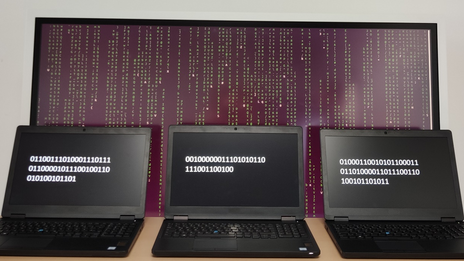 Three laptops standing next to each other with numeric codes, behind them a large screen with many characters one below the other.