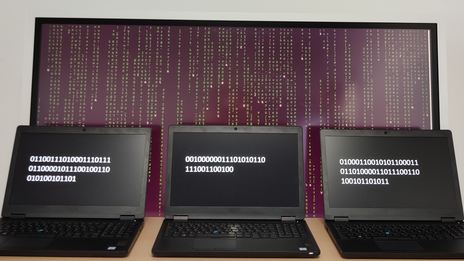 Drei nebeneinander stehende Laptops mit Zahlencodes, dahinter ein großer Bildschirm mit vielen Zeichen untereinander.