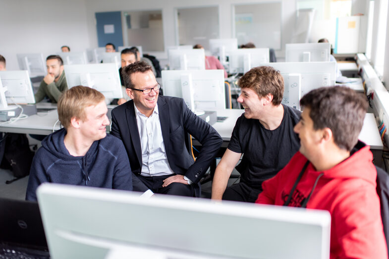 Photo of a lecturer sitting between students in the computer room. Everyone is looking at the professor with a smile.