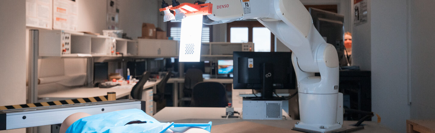 Photo of a surgical robot in action. In front of it lies an artificial torso with a blue surgical drape over it.