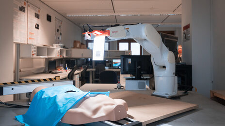 Photo of a surgical robot in action. In front of it lies an artificial torso with a blue surgical drape over it.