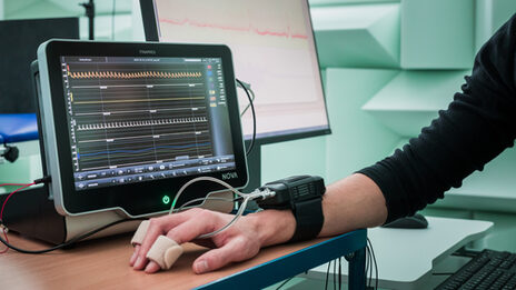 Photo of a hand with a connected measuring device on the table. Behind it, measurements are displayed or evaluated on a monitor.