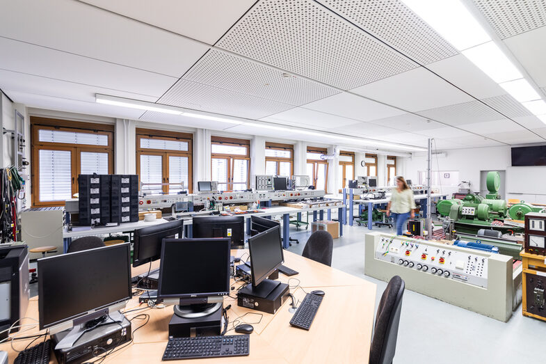 Photo of the laboratory for power electronics and drive systems. In the foreground is a computer desk, in the background a variety of laboratory equipment. A woman walks through the room.