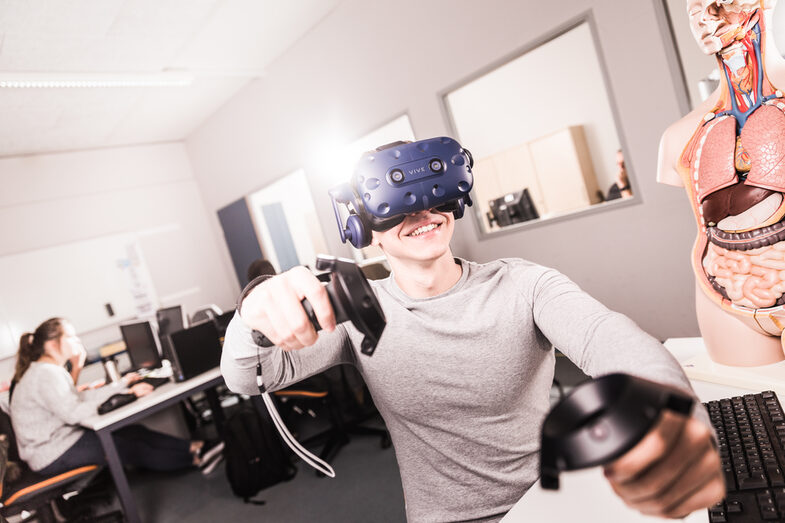 Foto von einem Studenten, der eine VR-Brille auf hat und Controller in der Hand hält, er sitzt an einem Rechnerarbeitsplatz. __A student has VR glasses on and controller in his hands,  is sitting at a computer workstation.